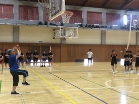 茶色いフローリングの体育館で、体操服のような青いTシャツと黒いハーフパンツ姿の指導者が、体育館の中央でバスケットボールのシュートフォームを実演し、その周りを取り囲むように多くの生徒たちが熱心に見学している様子の写真