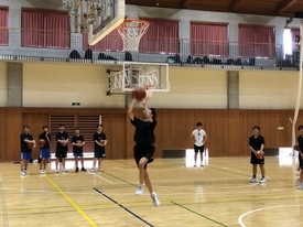 体育館の中で、数人の生徒がバスケットボールを持って並んで見守る中、一人の生徒がジャンプしてバスケットボールをゴールに向かってシュートしている写真
