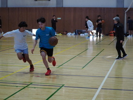 体育館のコート内で、青色のシャツを着てドリブルで切り込む選手と、それを阻止しようと白シャツの選手が並走して競り合う、緊迫したバスケットボールの練習の一場面を捉えた写真