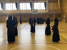 明るい体育館の中で防具と道着を身に纏った複数の剣士たちが稽古に励んでおり、手前では二人の剣士が竹刀を構えて向き合い、中央では指導者らしき人物が周囲を見守りながら立っている写真