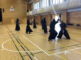 広々とした体育館で、剣道着と防具を身に纏った複数の生徒たちが二組に分かれて向かい合い、互いに竹刀を交えながら実戦形式の稽古に真剣な表情で取り組んでいる写真