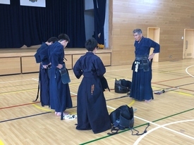 広々とした体育館で、剣道着を身に纏った三人の生徒たちが指導者と向き合い、稽古の合間に腰の防具を整えている写真