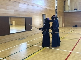 広々とした体育館で、剣道着を身に纏い竹刀を手に構え、指導者が後ろに立ち教えている写真