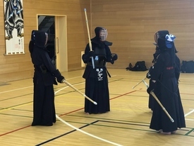 木目調の床が広がる屋内練習場で、防具を身に纏い竹刀を手にした剣道着姿の三人の人物が向き合い、中央の一人が何かを説明している写真