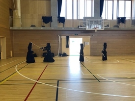 広々とした体育館で、防具を着用し竹刀を手にした剣道着姿の四人が、二組に分かれて間合いを取りながら稽古に励んでいる様子を遠目から捉えた写真