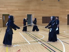 明るい体育館で剣道着と防具を身に纏った複数の剣士たちが稽古に励んでおり、手前では二人の剣士が互いに竹刀を交えて向き合い、その様子を右側から審判旗を持った指導者が静かに見守っている写真