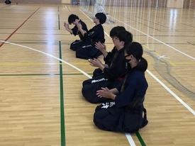 明るい体育館で剣道着と防具を身に纏った生徒たちが床に正座し、稽古に励む仲間たちに向けて真剣な表情で拍手を送りながら応援している写真