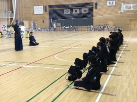 広々とした体育館で、剣道着と防具を身に纏った多くの生徒たちが床に一列に正座して整列し、前方で立ちながら話をする指導者の言葉を真剣な表情で静かに聞き入っている写真
