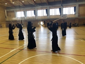 明るい体育館で防具を着用した複数の剣士が稽古に励んでおり、手前では二人の剣士が竹刀を高く振りかざして激しく打ち合い、奥では指導者がその様子を静かに見守っている写真