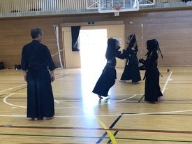 明るい体育館で剣道着と防具を身に纏った複数の剣士たちが稽古に励んでおり、手前では二人の剣士が互いに竹刀を振りかざして激しく打ち合い、その様子を左側から指導者が静かに見守っている写真