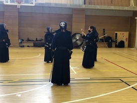 明るい体育館で剣道着と防具を身に纏った複数の剣士たちが稽古に励んでおり、中央では一人の剣士が周囲を見渡し、その右側では二人の剣士が向き合って竹刀を構えながら稽古の開始を待っている写真