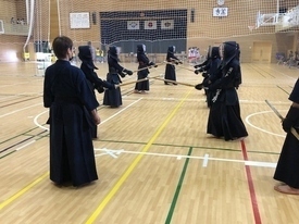 明るい体育館で剣道着と防具を身に纏った多くの剣士たちが一斉に稽古に励んでおり、手前では指導者が竹刀を手に持ちながら、向かい合って竹刀を交える生徒たちの様子を真剣な表情で見守っている写真