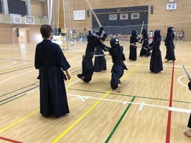 体育館で剣道着と防具を身に纏った多くの剣士たちが一斉に稽古に励んでおり、手前では指導者が竹刀を手に持ちながら、向かい合って竹刀を交える生徒たちの様子を見守っている写真