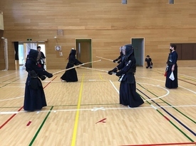 明るい体育館で剣道着と防具を身に纏った複数の剣士たちが稽古に励んでおり、手前では二人の剣士が互いに竹刀を交えて向き合い、その様子を右側から指導者が静かに見守っている