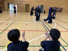 明るい体育館で剣道着と防具を身に纏った複数の剣士たちが稽古に励んでおり、手前では二人の人物が正座をして稽古の様子を見守りながら拍手を送っている写真