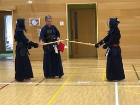 木目調の壁が印象的な体育館にて、指導者が竹刀に手を添えながら二人の剣士に向けて説明をしている写真