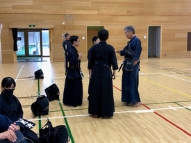 木目調の体育館にて、赤い審判旗を腰に差した指導者が二人の剣士に向けて熱心に身振り手振りで助言を行っている写真