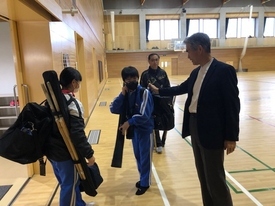 体育館で、青いジャージを着用し剣道防具袋や竹刀を背負った学生とに対し、スーツ姿の男性が笑顔で声を掛けている写真