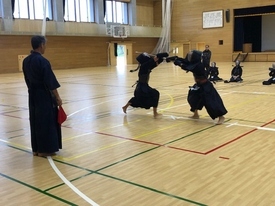 木目調の壁に囲まれた広い体育館で、手前で審判が見守る中、防具を身に着けた二人の剣士が互いに竹刀を高く振り上げて激しく打ち合っている写真