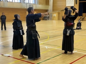 体育館で剣道着と防具を身に纏った三人の剣士が稽古に励んでおり、中央の年配の剣士が竹刀を真っ直ぐに突き出し、右側の剣士がそれを受けるように竹刀を頭上に掲げて構えている写真