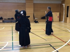 木目調の壁に囲まれた体育館にて、防具と面を装着し竹刀を手にした二人の剣士が、赤い旗を腰に差し指導を行う年配の男性から指導を受けている様子の写真