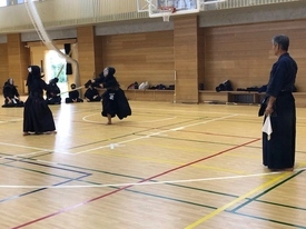 明るい照明の体育館にて、審判旗を手にした指導者が見守る中で二人の剣士が激しく竹刀を交えており、一方が踏み込んで面を打つ瞬間の躍動感と、周囲で見学する他の門下生たちの静かな緊張感が伝わってくる稽古風景の写真