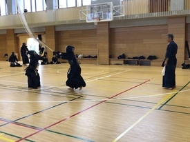 明るい照明の体育館にて二人の剣士が激しく竹刀を交えており、その後ろで指導者が見守っている写真