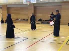 防具と面を装着し竹刀を手にした二人の剣士が、赤い旗を手に指導を行う男性の話を聞いている写真