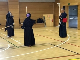 木目調の壁に囲まれた体育館にて、防具と面を装着し竹刀を手にした二人の剣士が、赤い旗を手に持つ指導の説明に耳を傾けている様子の写真