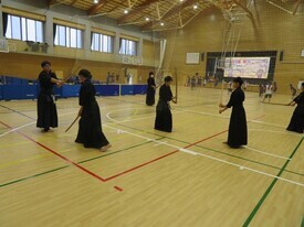 広い体育館のコート内で複数の剣士たちが竹刀を構えて稽古に励み、手前では二人の競技者が互いに間合いを測りながら向き合っている写真
