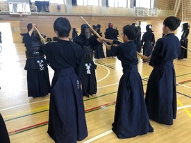 明るい光が差し込む体育館で、手前には紺色の道着を纏い竹刀を構える背中姿の生徒たちが、奥には防具をフル装備して稽古相手を務める生徒たちが対峙し、互いに真剣な表情で打ち込みの練習を行っている写真