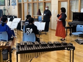 木目調の室内で手前に置かれたシロフォンの奥に、譜面台に向かって座る生徒たちとそれを見守る大人二人が立っており、ピアノやメトロノームが置かれた音楽室のような場所で練習を行う吹奏楽部の様子を写した写真