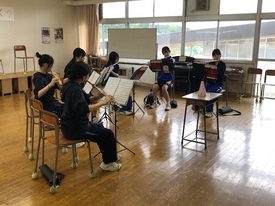 明るい光が差し込む窓際の練習場で、フルートを構えた生徒たちが譜面台を囲むように座り、メトロノームが置かれた教卓を囲んで集中した様子でパート練習に取り組んでいる吹奏楽部の活動風景を捉えた写真