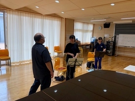 木目調の広い練習室で楽器を手にした二人の女子生徒が、手前に立つ男性指導者のアドバイスを真剣な表情で聞いている写真