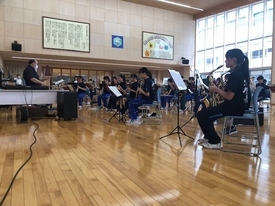 木目調の広いホールで、サックスやクラリネットなどの楽器を構えた吹奏楽部の生徒たちが譜面台を前に整列して座り、左端でタクトを振る男性指揮者の合図に合わせて真剣な表情で合奏練習に取り組んでいる写真