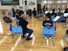 木目調の床が広がる室内で、トロンボーンを構えた指導者らしき男性がトロンボーンを構えた生徒に、指導している様子を後ろから捉えた写真