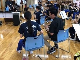 木目調の広い室内でサックスを構えた中学生たちが青い椅子に座り、指導者と思われる男性と共に真剣な表情で合奏練習に取り組んでいる、活気ある吹奏楽部の活動風景を背後から捉えた写真