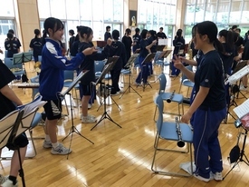 明るい日差しが差し込む広い練習場で、青いジャージや黒いTシャツ姿の生徒たちが楽器を椅子に置き、笑顔で談笑したり手指の運動をしたりしてリラックスした時間を過ごしている、吹奏楽部の練習の合間の風景を捉えた写真