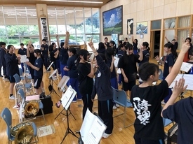 明るい練習場で青いジャージや黒いTシャツ姿の生徒たちが一斉に片手を高く突き上げ、笑顔でポーズを取ったり談笑したりして一体感を楽しんでいる、吹奏楽部の活気あふれる練習風景を捉えた写真