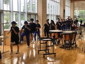 明るい光が差し込む広い練習場で、黒いTシャツや青いジャージを着用した吹奏楽部の生徒たちが、ティンパニなどの打楽器が並ぶ横で一列になって足踏みを行っている様子の写真