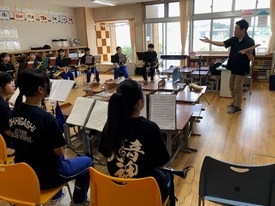 木目調の教室で黒いTシャツや青いジャージを着用した吹奏楽部の生徒たちが、譜面台を前に椅子に座り、身振り手振りを交えて熱心に指導する男性の話を真剣な表情で聞いている、練習風景を背後から捉えた写真