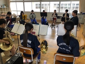 明るい光が差し込む窓際の練習場で、ホルンやユーフォニアムなどの金管楽器を手にした生徒たちが、譜面台を囲むように円形に並んで座り、集中した様子で合同練習に取り組んでいる吹奏楽部の活動風景を背後から捉えた写真