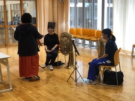 明るい光が差し込む木目調の練習場で、チューバなどの大型楽器を置いた二人の女子生徒が椅子に座り、ロングスカートを履いた女性指導者の話を笑顔で聞いている写真