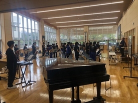 陽光が差し込む広々とした音楽室で、手前に置かれたグランドピアノの奥に大勢の部員たちが集まり、右前方に立つ指導者のもとで合奏や練習の準備を行っている、活気あふれる吹奏楽部の活動風景の写真