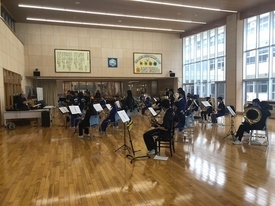 陽光が差し込む広いホールで、青いジャージや黒い服を着た吹奏楽部員たちが、一定の間隔を保ちながら椅子に座り、譜面台の楽譜を真剣な表情で確認しながら各々の管楽器で合奏練習に取り組んでいる活気ある風景の写真