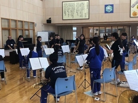 木目調の広い室内で、黒いTシャツや青いジャージを着用した中学生たちが楽器を手に持ち向き合っている写真