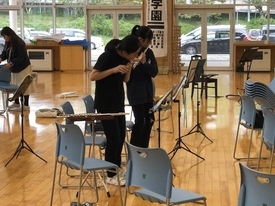 明るい室内で黒いTシャツや青いジャージを着た二人の生徒が、譜面台の前に立って楽譜を確認しながら演奏する練習風景の写真