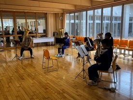 窓から明るい陽光が差し込む広々とした音楽室内で、大きなチューバやユーフォニアムを構えた吹奏楽部員たちが、一定の間隔を空けて椅子に座り、譜面台の楽譜を確認しながら低音パートの個人練習や音出しを行っている写真