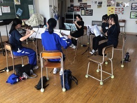 木目調の床が広がる明るい室内で、黒いTシャツや青いジャージを着用した生徒たちが円状に椅子を並べて座り、譜面台に置かれた楽譜を真剣に見つめながらフルートなどの木管楽器のパート練習に集中して取り組んでいる風景の写真