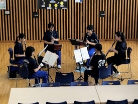 木目調の床が広がる明るい室内で、黒いTシャツを着用した5人の生徒たちが円状に椅子を並べて座り、譜面台に置かれた楽譜を真剣に見つめながらクラリネットのパート練習に集中して取り組んでいる風景の写真